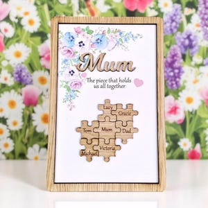 May include: Wooden framed artwork with the word "Mum" in cursive, floral accents, and a puzzle design with names. The text reads "The piece that holds us all together." The puzzle pieces include the names: Lucy, Gracie, Tom, Mum, Dad, Michael, and Victoria.