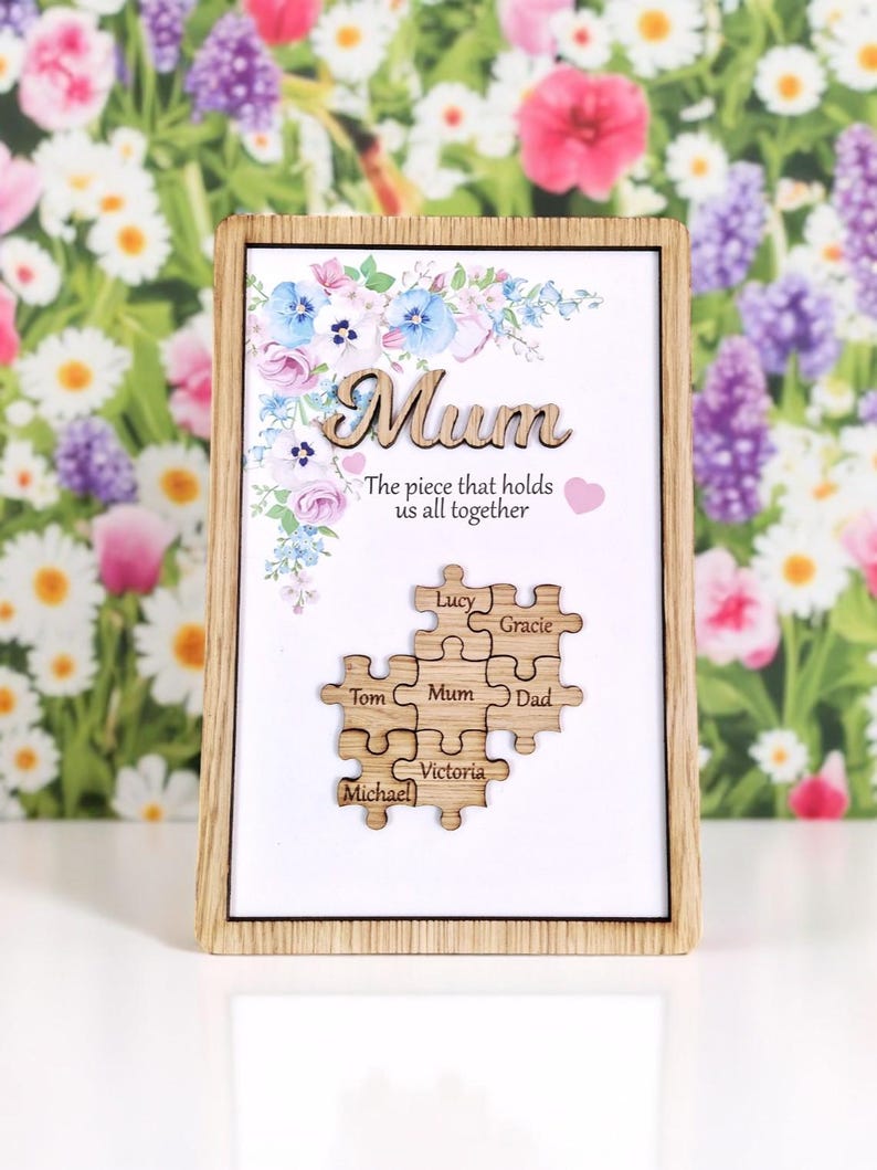 May include: Wooden framed artwork with the word "Mum" in cursive, floral accents, and a puzzle design with names. The text reads "The piece that holds us all together." The puzzle pieces include the names: Lucy, Gracie, Tom, Mum, Dad, Michael, and Victoria.