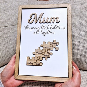 May include: Wooden framed sign with the word "Mum" in a cursive font. The text "the piece that holds us all together" is below. Puzzle pieces with names are below the text.
