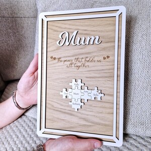 May include: Wooden framed plaque with the word "Mum" in cursive. The plaque has a light wood grain background and a white border. It features puzzle pieces with names, and the phrase "the piece that holds us all together".