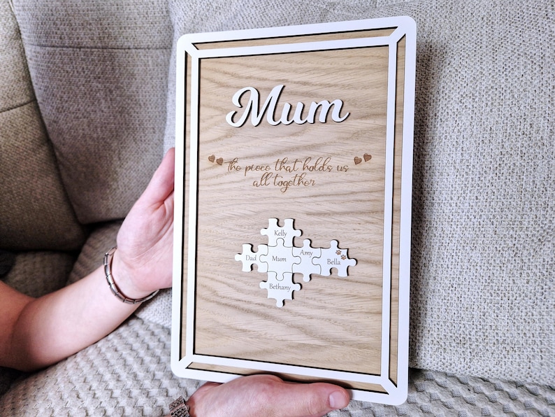 May include: Wooden framed plaque with the word "Mum" in cursive. The plaque has a light wood grain background and a white border. It features puzzle pieces with names, and the phrase "the piece that holds us all together".