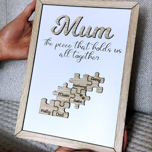 May include: A framed wooden sign with the word "Mum" in a cutout design. The sign includes the phrase "the piece that holds us all together" and puzzle piece cutouts with names. The frame is light brown.
