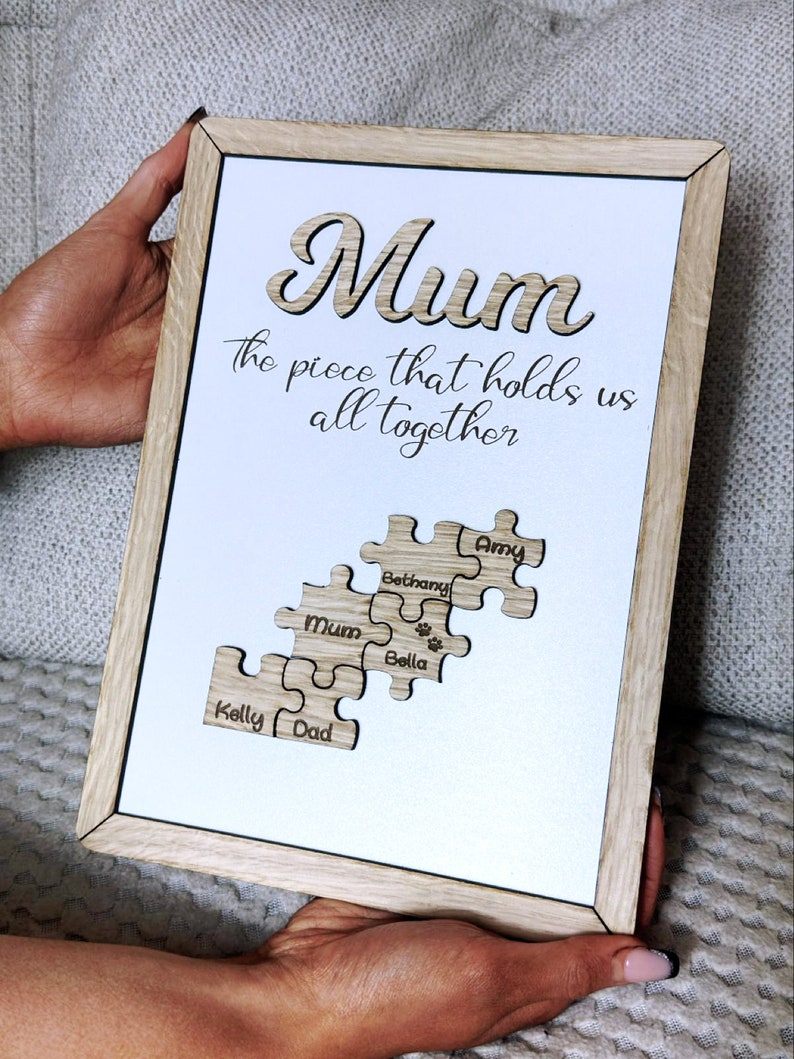 May include: A framed wooden sign with the word "Mum" in a cutout design. The sign includes the phrase "the piece that holds us all together" and puzzle piece cutouts with names. The frame is light brown.