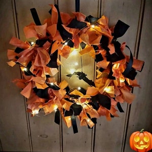 May include: A Halloween wreath made of orange and black fabric scraps with white fairy lights. The wreath is hanging on a wooden wall. A carved pumpkin is in the bottom right corner of the image.