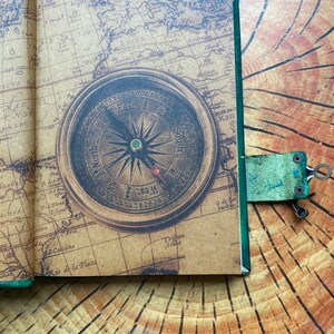 May include: Open journal with a vintage map design and a compass illustration. The journal is open to a page with a detailed map and a compass rose. A green leather bookmark with a metal clip is attached.