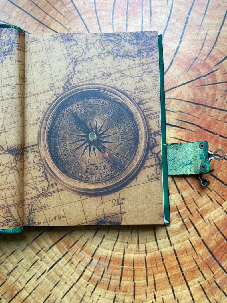 May include: Open journal with a vintage map design and a compass illustration. The journal is open to a page with a detailed map and a compass rose. A green leather bookmark with a metal clip is attached.