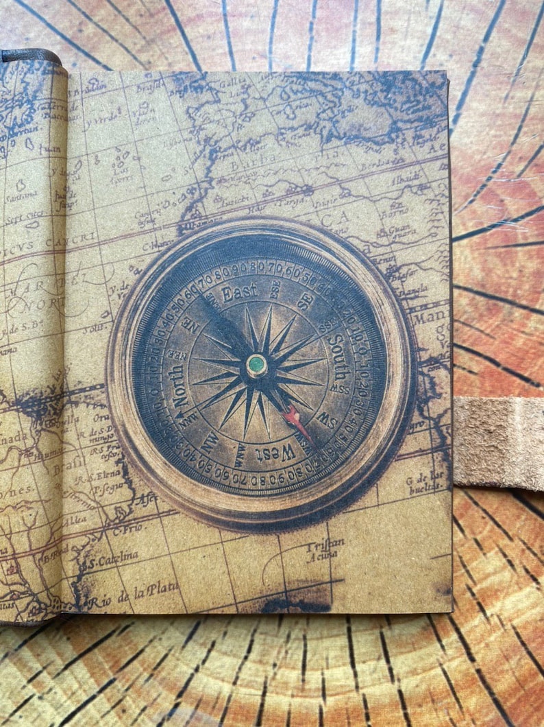 K&ouml;nnte beinhalten: Ein Kompass im Vintage-Stil mit braunem Lederbezug und einem Kartenhintergrund. Das Kompassgesicht ist mit den Himmelsrichtungen gekennzeichnet und hat eine goldfarbene Nadel.