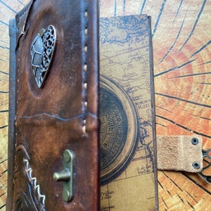 K&ouml;nnte beinhalten: Ein braunes, ledergebundenes Tagebuch mit einem Kompass-Design auf der Innenseite des Deckels. Das Tagebuch hat einen herzf&ouml;rmigen Metallapplikation auf dem vorderen Deckel und einen Lederriemen mit einem Metallverschluss.