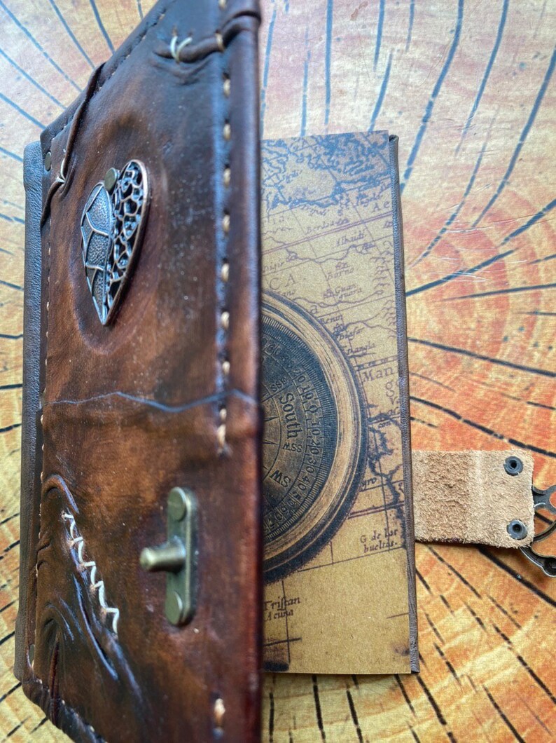 K&ouml;nnte beinhalten: Ein braunes, ledergebundenes Tagebuch mit einem Kompass-Design auf der Innenseite des Deckels. Das Tagebuch hat einen herzf&ouml;rmigen Metallapplikation auf dem vorderen Deckel und einen Lederriemen mit einem Metallverschluss.