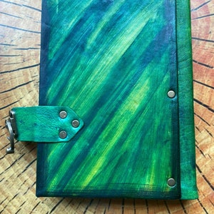 May include: A handmade journal with a vibrant green and blue abstract painted cover. The leather-bound journal features a flap closure with a metal clasp and is secured with metal rivets. The journal is displayed on a wooden surface.
