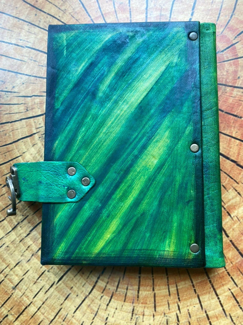 May include: A handmade journal with a vibrant green and blue abstract painted cover. The leather-bound journal features a flap closure with a metal clasp and is secured with metal rivets. The journal is displayed on a wooden surface.