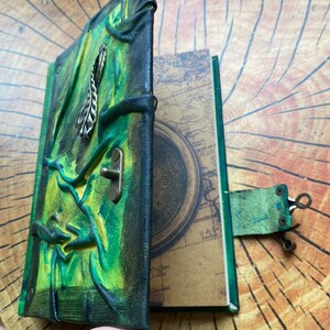 May include: A handmade leather journal with a green and black cover. The cover features an embossed design and a metal clasp. The pages inside have a map design. The journal is open, revealing the interior pages.