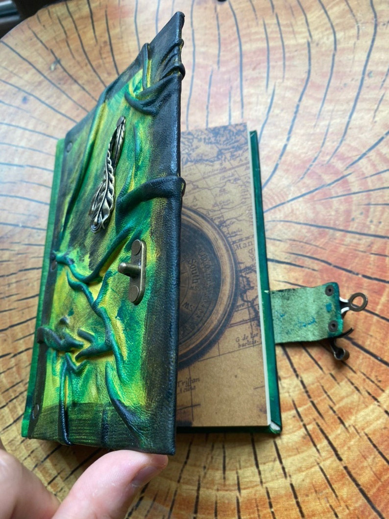 May include: A handmade leather journal with a green and black cover. The cover features an embossed design and a metal clasp. The pages inside have a map design. The journal is open, revealing the interior pages.