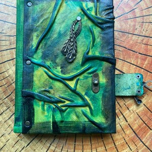 May include: Hand-painted leather journal with a green and yellow color scheme. The cover features embossed tree branch designs and a metal feather charm. A leather strap with a metal clasp secures the journal.