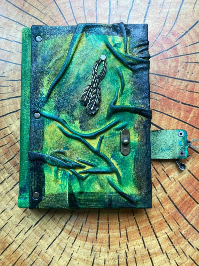 May include: Hand-painted leather journal with a green and yellow color scheme. The cover features embossed tree branch designs and a metal feather charm. A leather strap with a metal clasp secures the journal.