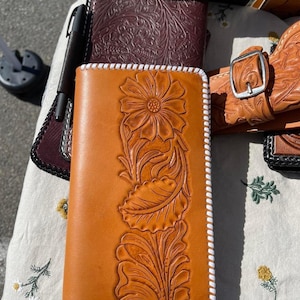 May include: A brown leather wallet with a white stitched border. The wallet has a floral design carved into the leather.