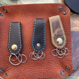 May include: Three leather keyrings with metal key rings. The keychains are brown, black, and black with a blue stitch. The keychains are on a brown leather surface.