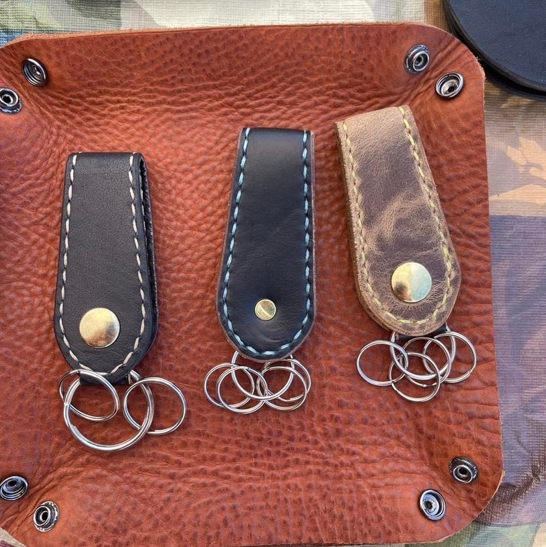 May include: Three leather keyrings with metal key rings. The keychains are brown, black, and black with a blue stitch. The keychains are on a brown leather surface.