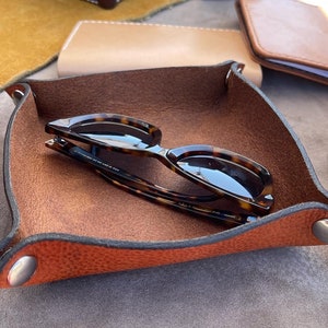 May include: A brown leather tray with a dark brown suede interior. The tray has two silver metal snaps and a pair of tortoiseshell sunglasses are resting inside.