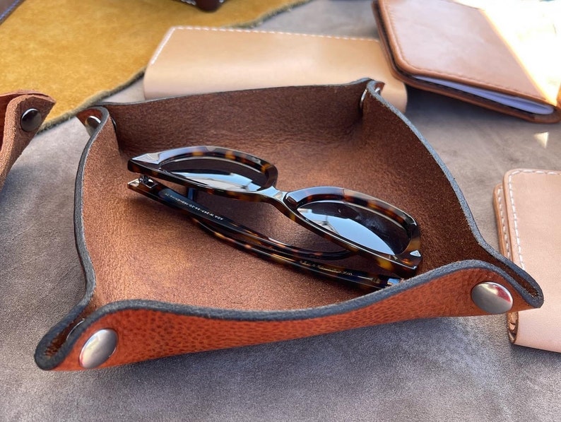 May include: A brown leather tray with a dark brown suede interior. The tray has two silver metal snaps and a pair of tortoiseshell sunglasses are resting inside.