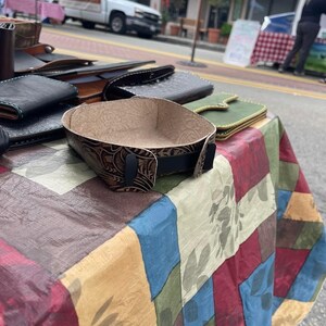 May include: A brown leather tray with a tooled design sits on a colourful patterned tablecloth. The tray is empty and ready to hold small items.