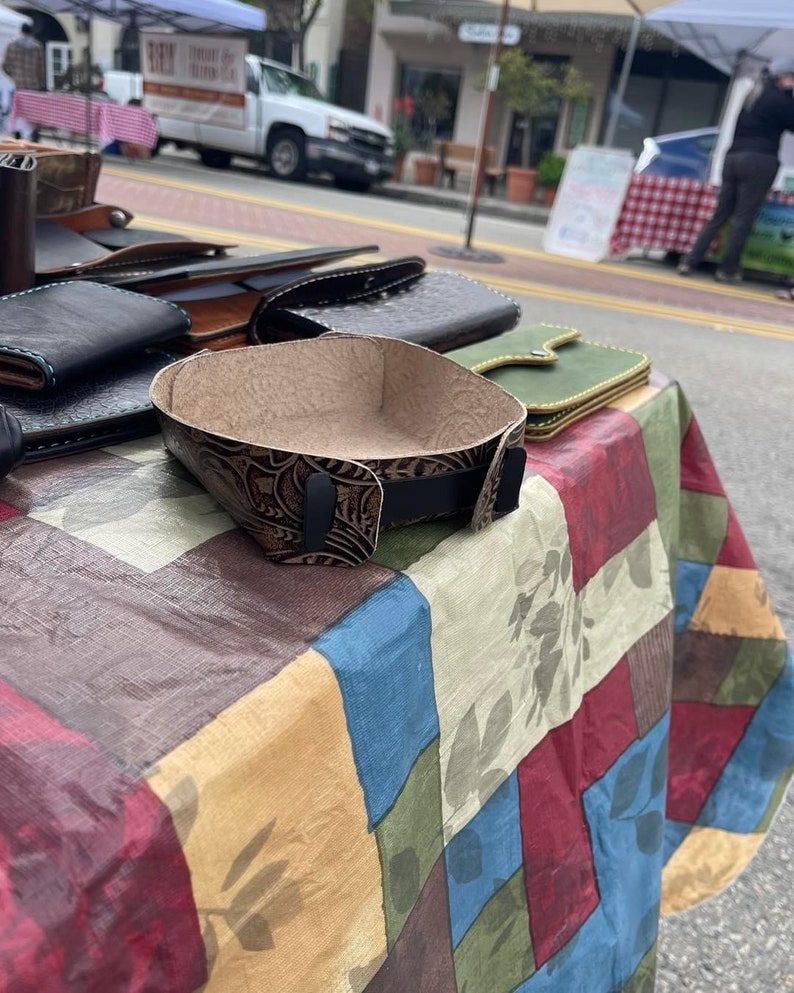 May include: A brown leather tray with a tooled design sits on a colourful patterned tablecloth. The tray is empty and ready to hold small items.