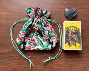 May include: A green drawstring bag with a tropical toucan print. The bag is tied with a green cord and has a silver charm. The bag is sitting on a wooden surface next to a tarot card with the number IX and a green heart-shaped stone.