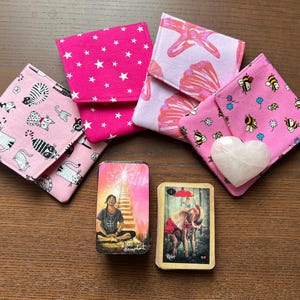 May include: A collection of pink fabric items, including small square pouches with patterns of white stars, cats, bees, and seashell designs. Two tarot card decks and a heart-shaped stone are also present.