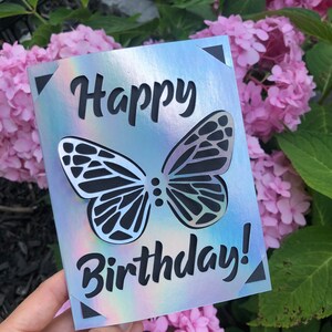 May include: A holographic birthday card with a butterfly design. The card features the words "Happy Birthday!" cut out in black. The butterfly is also cut out, revealing the background.