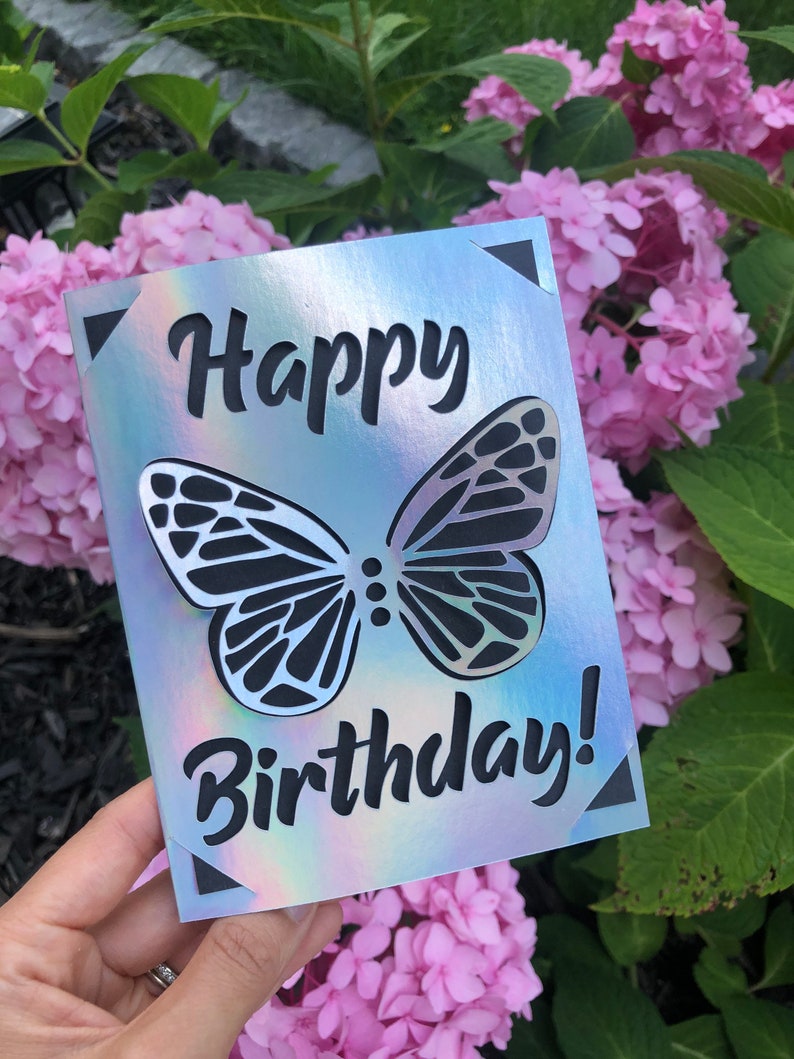 May include: A holographic birthday card with a butterfly design. The card features the words "Happy Birthday!" cut out in black. The butterfly is also cut out, revealing the background.