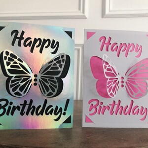 May include: Two birthday cards with butterfly cutouts. One card has a holographic background with a black butterfly and text. The other card has a white background with a pink butterfly and text. The words "Happy Birthday!" are cut out.