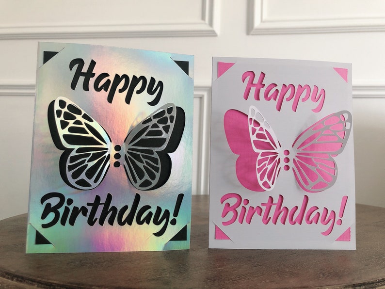 May include: Two birthday cards with butterfly cutouts. One card has a holographic background with a black butterfly and text. The other card has a white background with a pink butterfly and text. The words "Happy Birthday!" are cut out.
