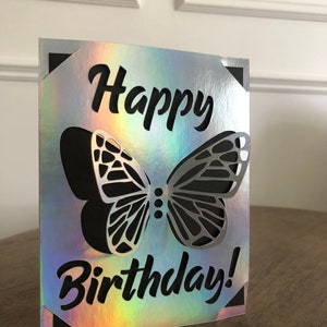 May include: A silver and iridescent birthday card with a cut-out butterfly design. The words "Happy Birthday!" are written in black script. The card has black corner accents.