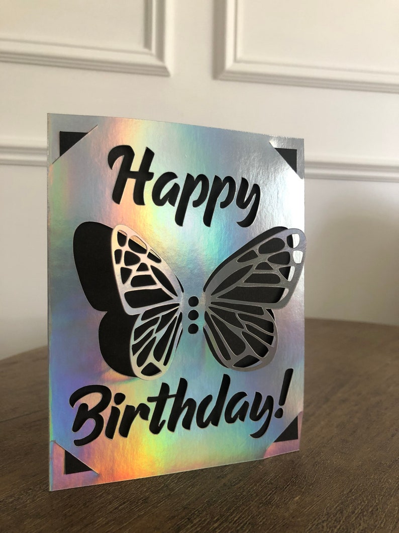 May include: A silver and iridescent birthday card with a cut-out butterfly design. The words "Happy Birthday!" are written in black script. The card has black corner accents.
