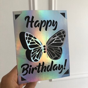May include: A holographic birthday card with a cut-out butterfly design. The words "Happy Birthday!" are in black script. The card has a reflective, iridescent finish, with a black butterfly silhouette.