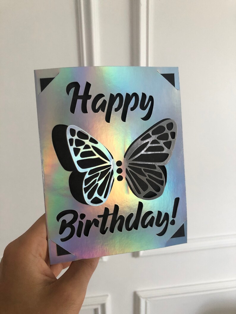 May include: A holographic birthday card with a cut-out butterfly design. The words "Happy Birthday!" are in black script. The card has a reflective, iridescent finish, with a black butterfly silhouette.