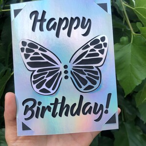 May include: A birthday card with a holographic finish. The card features a cut-out butterfly design and the words "Happy Birthday!" in a bold, black font. The corners of the card have black triangle accents.