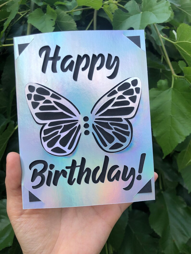May include: A birthday card with a holographic finish. The card features a cut-out butterfly design and the words "Happy Birthday!" in a bold, black font. The corners of the card have black triangle accents.