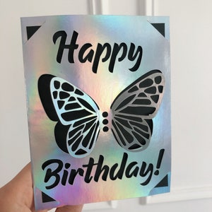 May include: A holographic birthday card with a butterfly design. The card features a cut-out butterfly with black and silver wings, and the words "Happy Birthday!" in black script. The card has black corner accents.