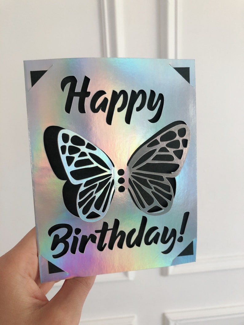 May include: A holographic birthday card with a butterfly design. The card features a cut-out butterfly with black and silver wings, and the words "Happy Birthday!" in black script. The card has black corner accents.