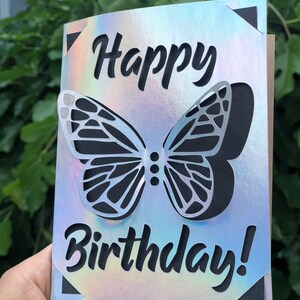 May include: A birthday card with a holographic finish. The card features a cut-out butterfly design and the words "Happy Birthday!" in black script. The corners of the card have black triangle accents.