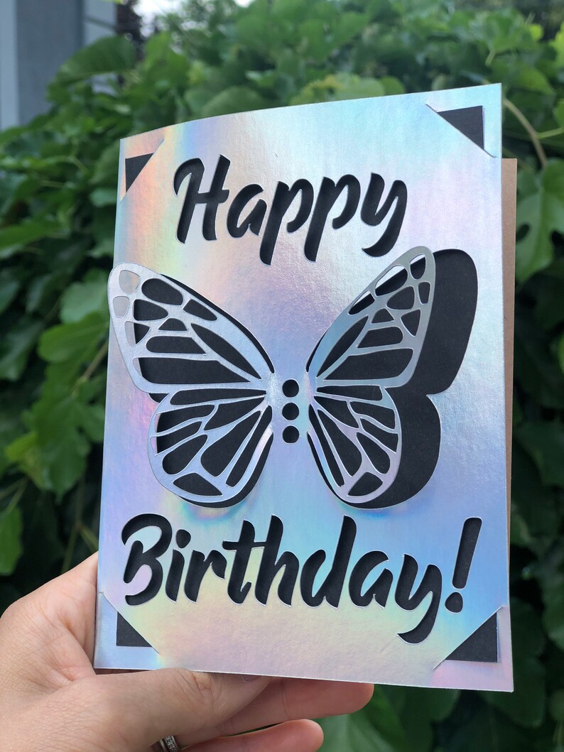 May include: A birthday card with a holographic finish. The card features a cut-out butterfly design and the words "Happy Birthday!" in black script. The corners of the card have black triangle accents.