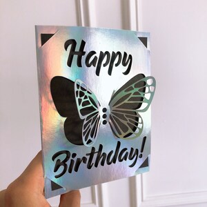 May include: A holographic birthday card with a cut-out butterfly design. The words "Happy Birthday!" are cut out in a bold, black font. The card has a reflective, iridescent finish, with black corner accents.