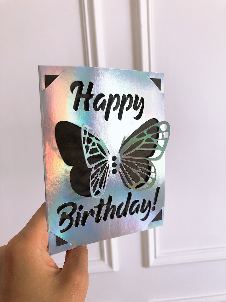 May include: A holographic birthday card with a cut-out butterfly design. The words "Happy Birthday!" are cut out in a bold, black font. The card has a reflective, iridescent finish, with black corner accents.