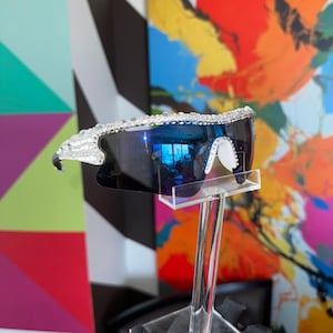 Pu&ograve; includere: Un paio di occhiali da sole blu a specchio con montatura in strass trasparente. Gli occhiali da sole sono esposti su un supporto trasparente su uno sfondo colorato.