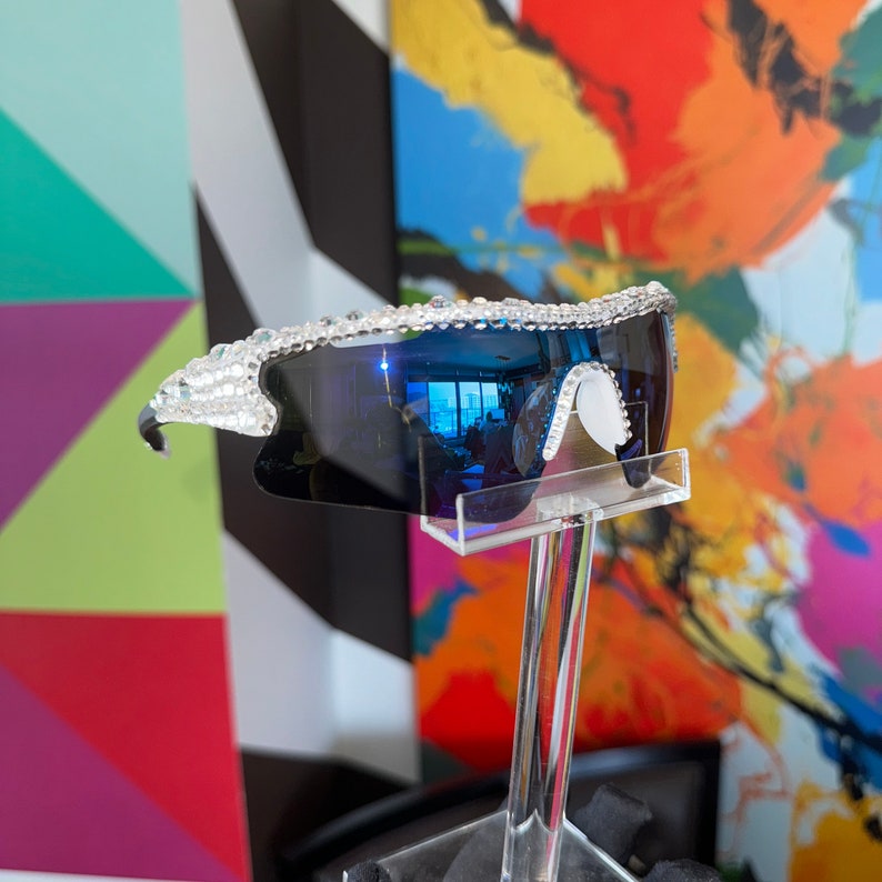 Pu&ograve; includere: Un paio di occhiali da sole blu a specchio con montatura in strass trasparente. Gli occhiali da sole sono esposti su un supporto trasparente su uno sfondo colorato.