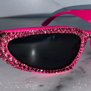 May include: Hot pink sunglasses with black lenses. The frames are embellished with pink rhinestones. The sunglasses have a cat-eye shape and are set against a light background.