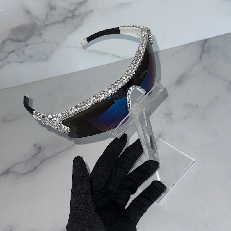 Pu&ograve; includere: Un paio di occhiali da sole con montatura argentata e lenti blu. La montatura &egrave; ricoperta di strass.