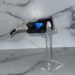 Pu&ograve; includere: Un paio di occhiali da sole neri con lenti a specchio blu e una bordatura in strass argentati. Gli occhiali da sole sono esposti su un supporto in acrilico trasparente.