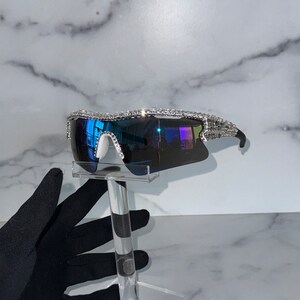 Pu&ograve; includere: Un paio di occhiali da sole con montatura argentata e lenti blu. La montatura &egrave; decorata con strass.